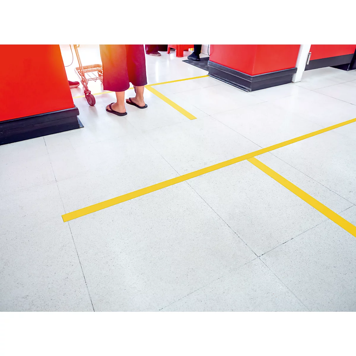 Person in red trousers waiting at the checkout. Yellow lines mark the distance on white floor.