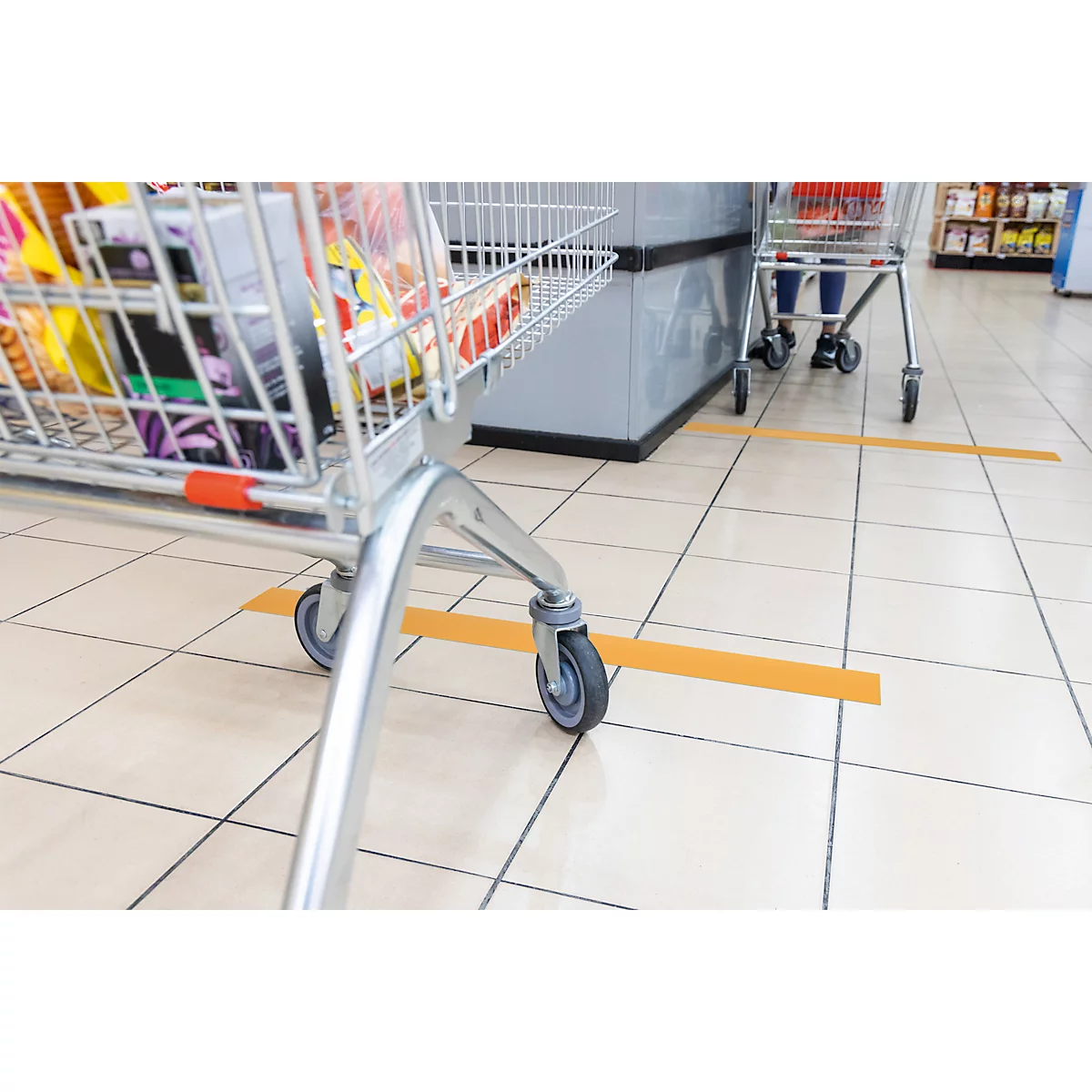 Shopping cart filled with groceries stands in front of a checkout counter. Yellow line on the floor marks distance.