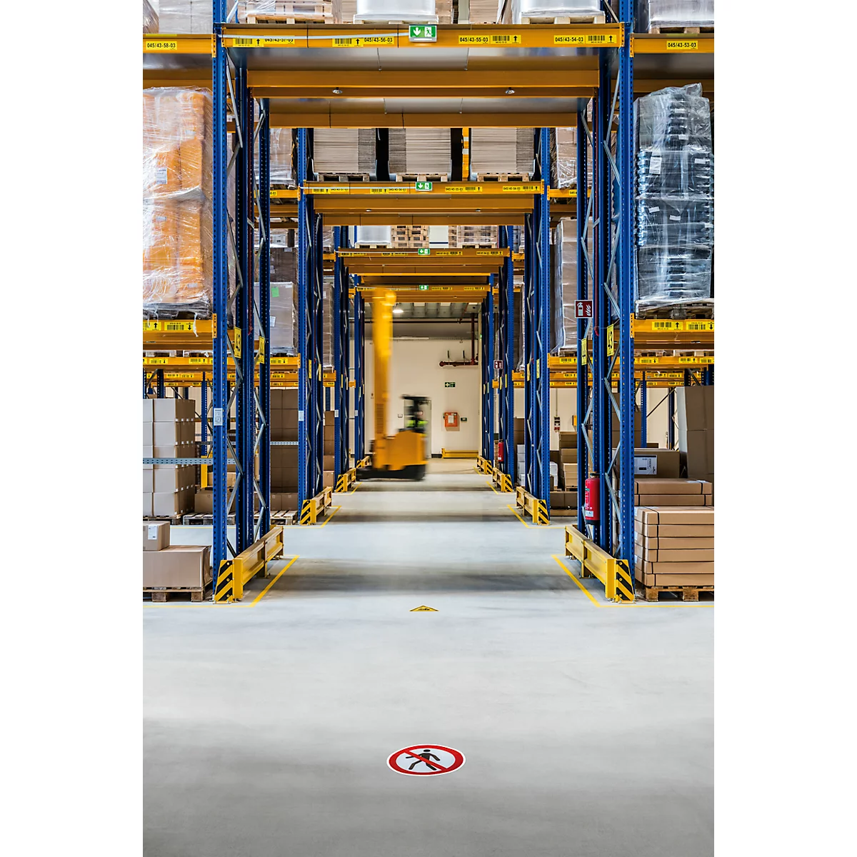 Warehouse with high shelves. A forklift truck drives in the distance. A sign with a prohibited footpath is on the floor.