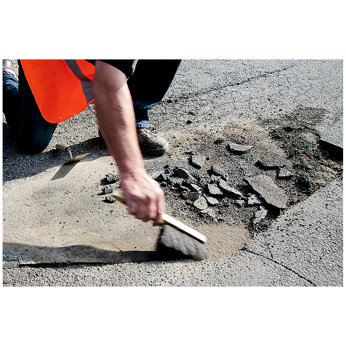 A person sweeps a road that has a pothole. The worker wears an orange safety vest.