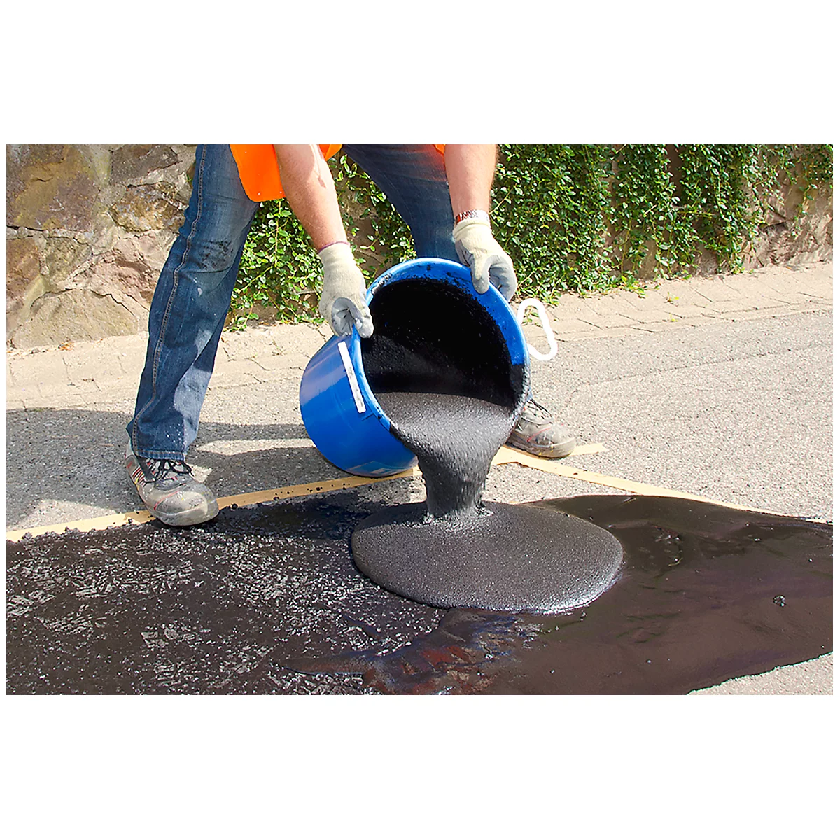Person pours black asphalt from blue bucket onto street. Work clothes, gloves.