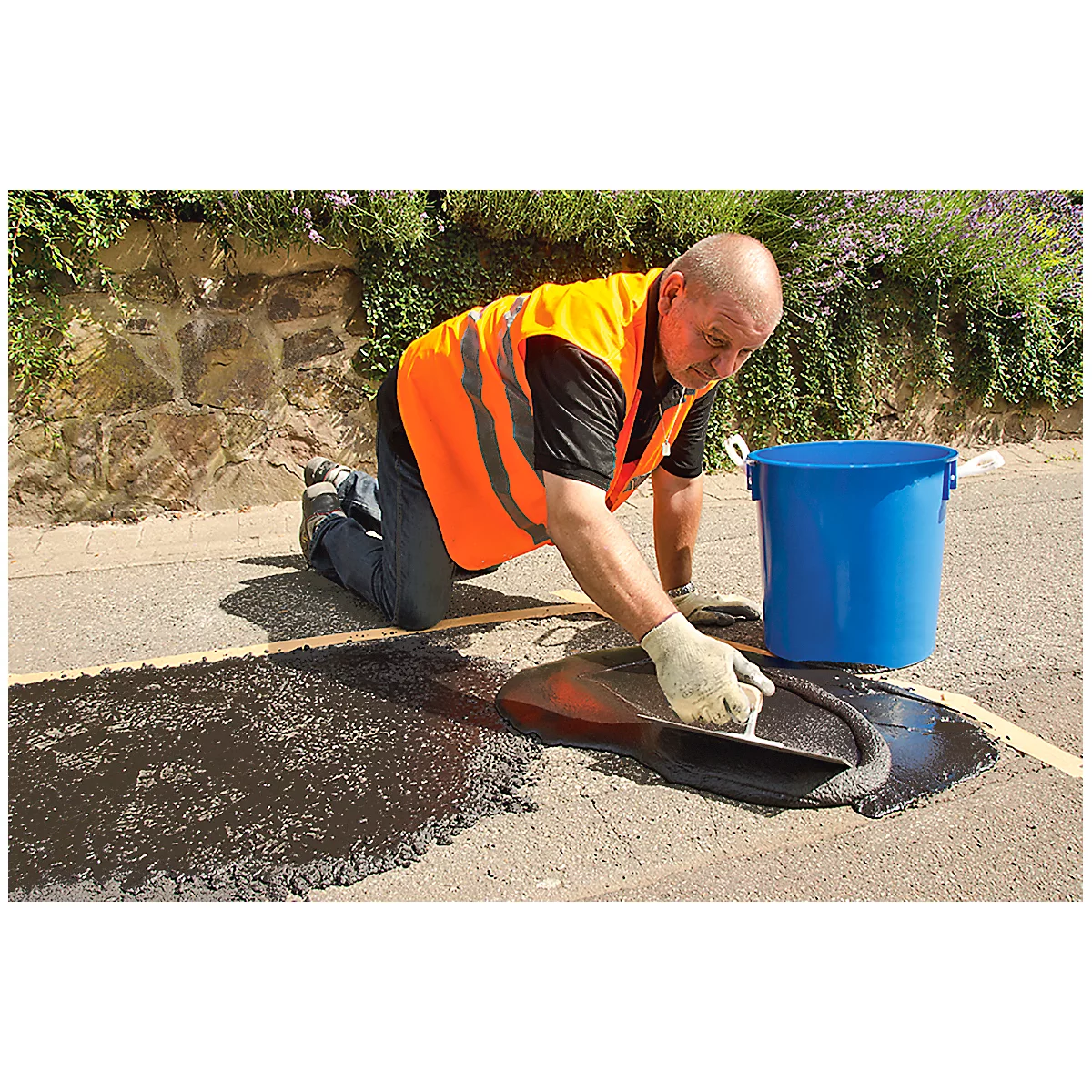 A man kneels on the road, repairing an asphalt hole with a trowel. He wears a safety vest and gloves, and a blue bin stands next to him.