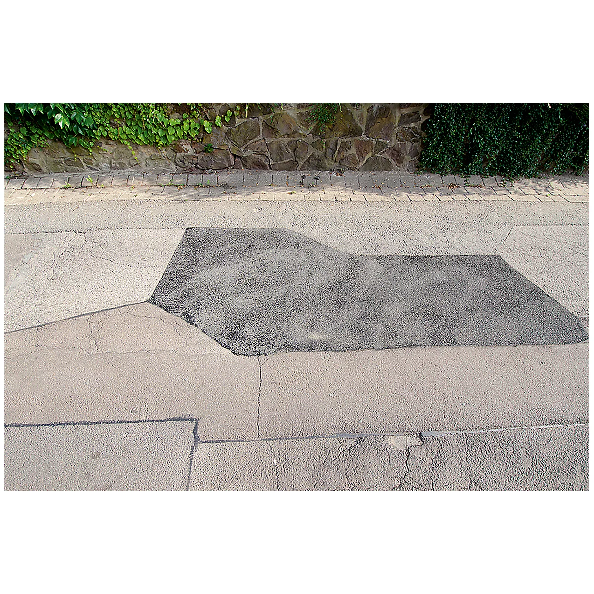 Patch of asphalt on a road, surrounded by gray paving. Top: wall with greenery. Top view.