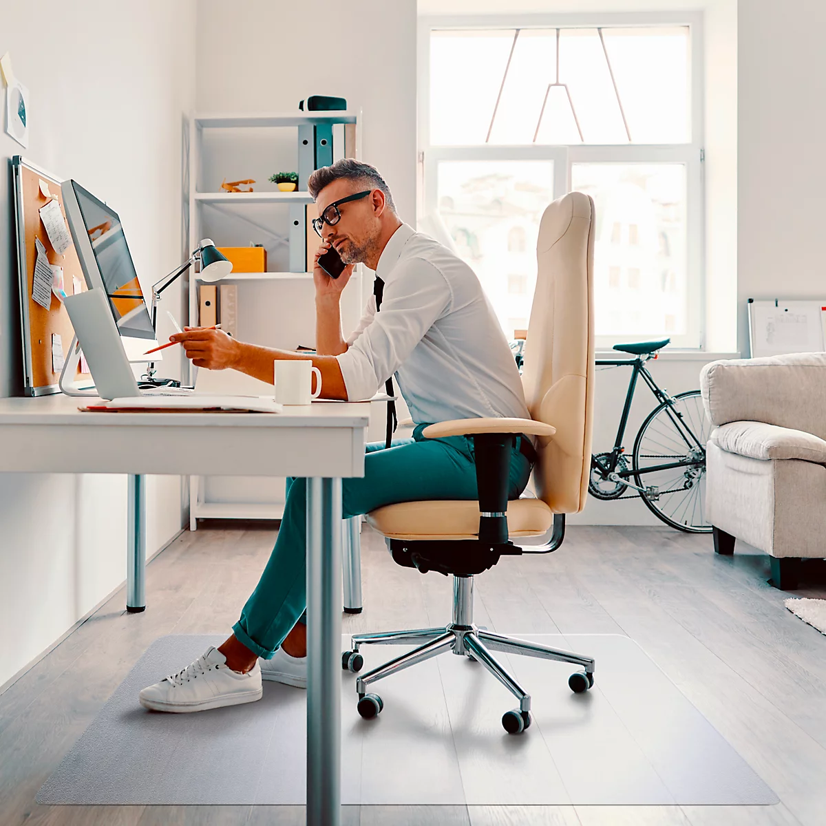 Een man in een shirt telefoneert en wijst naar een laptop. Hij zit aan een bureau met een stoel in een helder kantoor.
