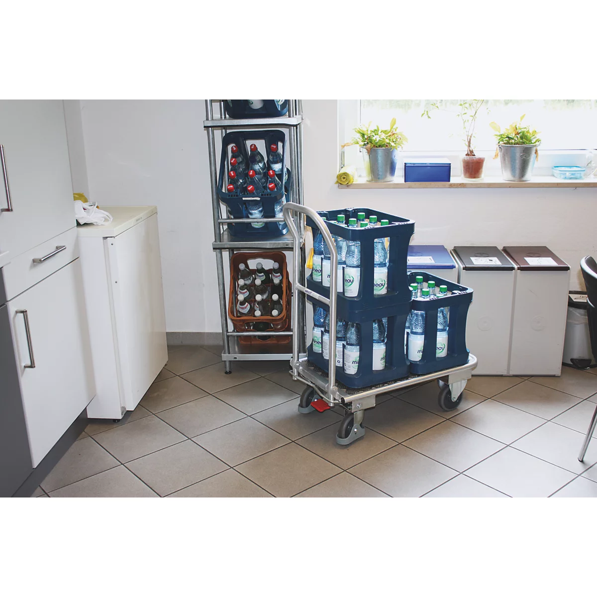 A serving cart with beverage crates in a room. Shelves and a window with plants can be seen in the background.