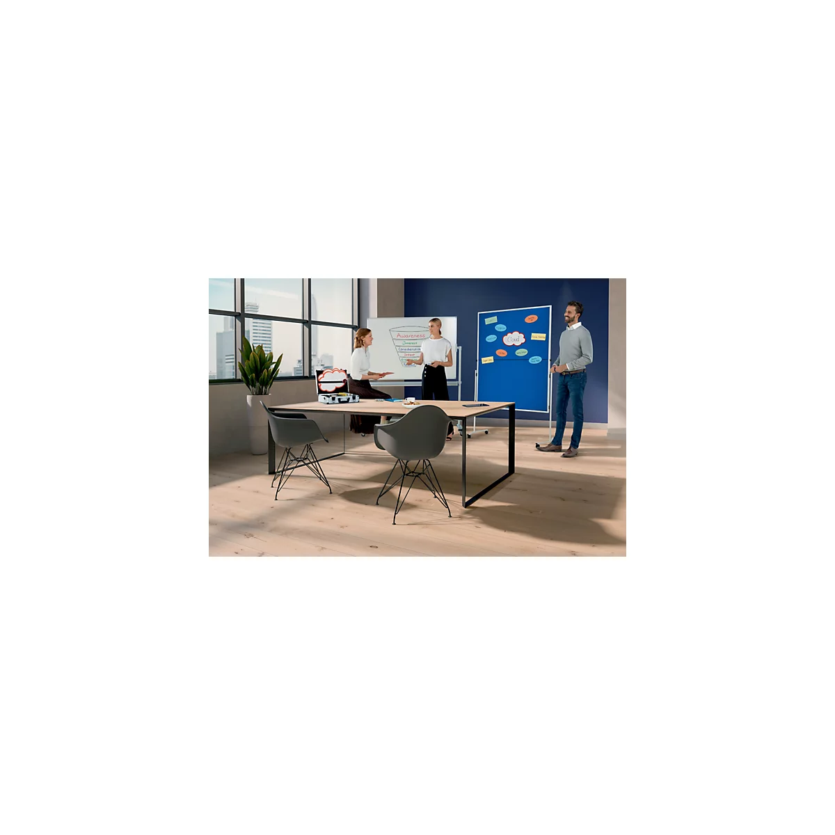 Drie mensen in een kantoor die brainstormen aan een tafel met twee stoelen. Op de achtergrond een whiteboard en een prikbord.