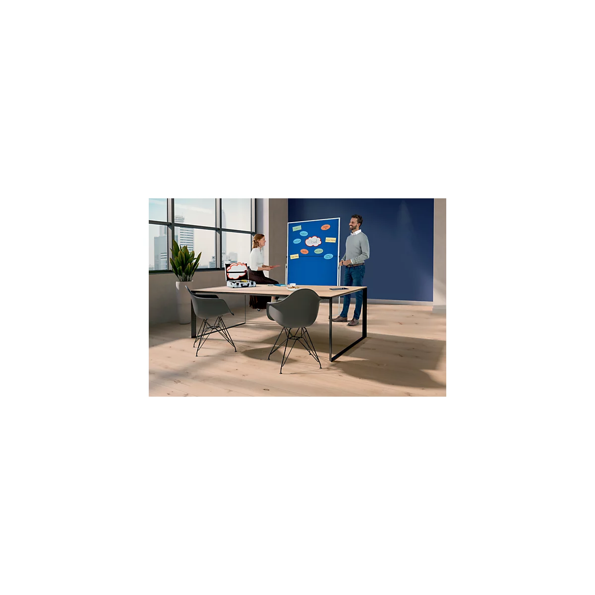 Twee mensen staan voor een blauw prikbord. Er staat een doos op de tafel en twee stoelen ervoor.