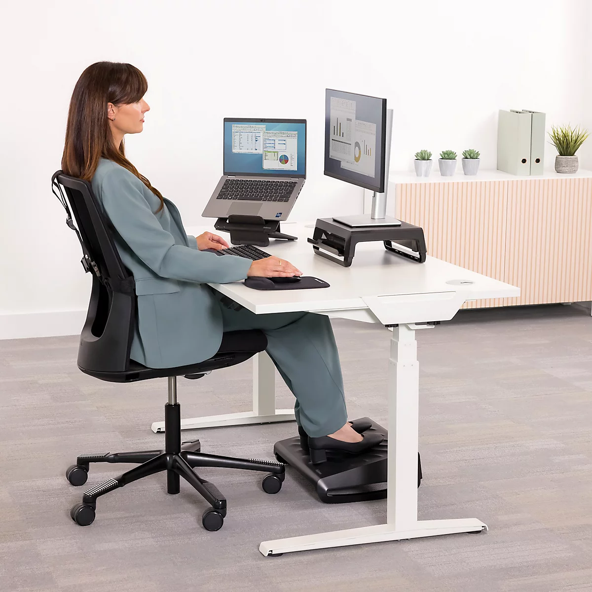 Eine Frau sitzt an einem weißen höhenverstellbaren Schreibtisch und arbeitet mit einem Laptop und einem Monitor. Sie hat eine ergonomische Körperhaltung.