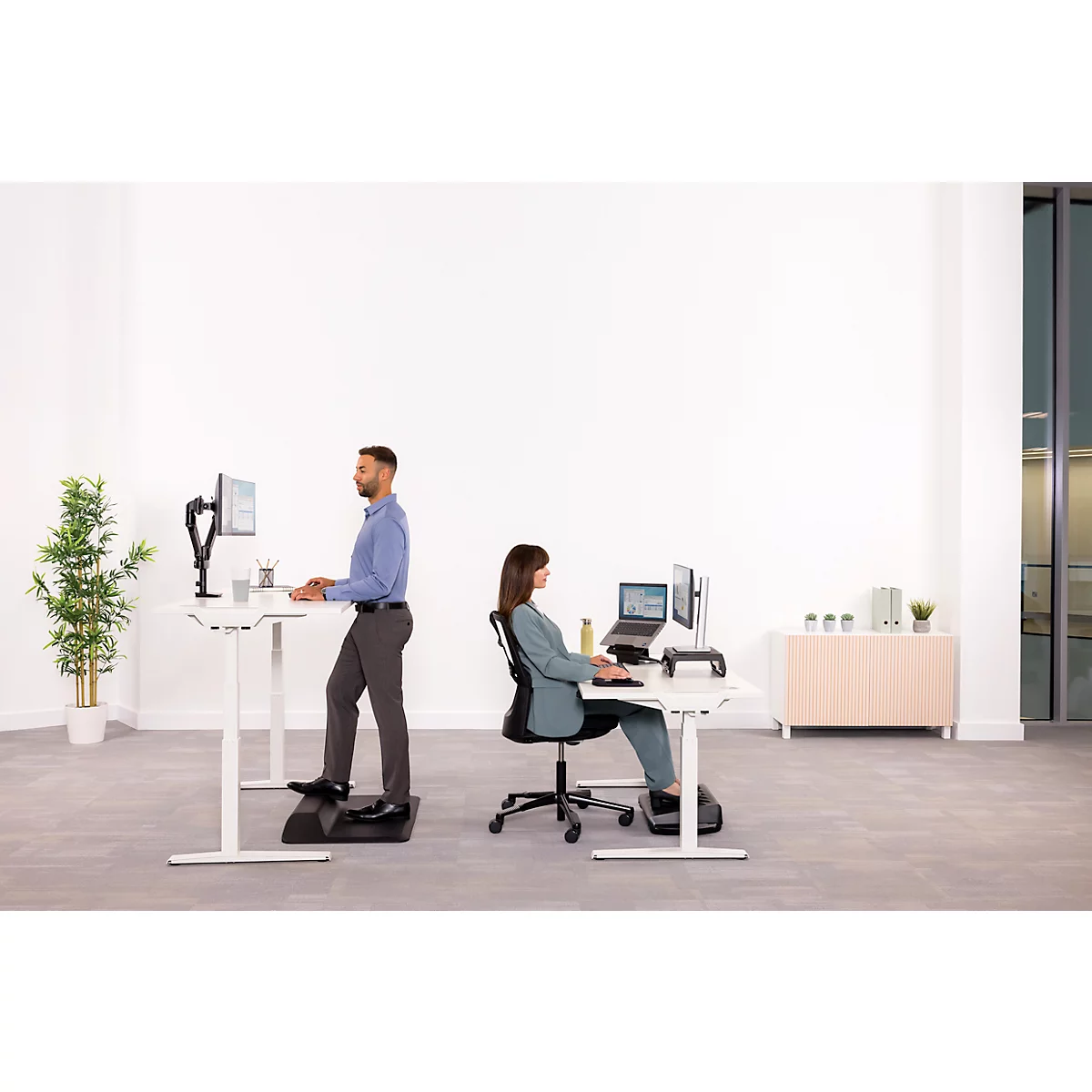 Ein Mann steht an einem höhenverstellbaren Schreibtisch, eine Frau sitzt. Beide arbeiten an Computern in einem hellen Büro.