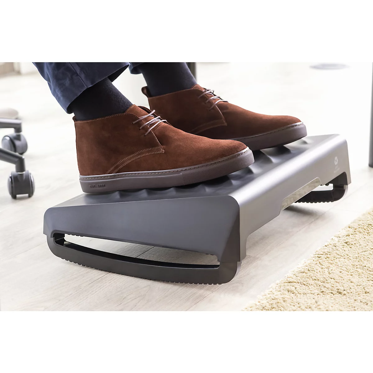 Füße in braunen Schuhen auf einer grauen Fußstütze. Die Füße ruhen auf einer Stütze, die unter einem Schreibtisch steht.