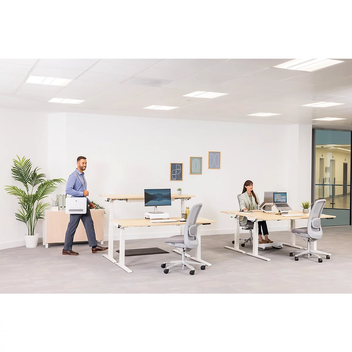 Büro mit zwei höhenverstellbaren Schreibtischen und Mitarbeitern. Ein Mann geht, eine Frau sitzt. Helle, moderne Atmosphäre.
