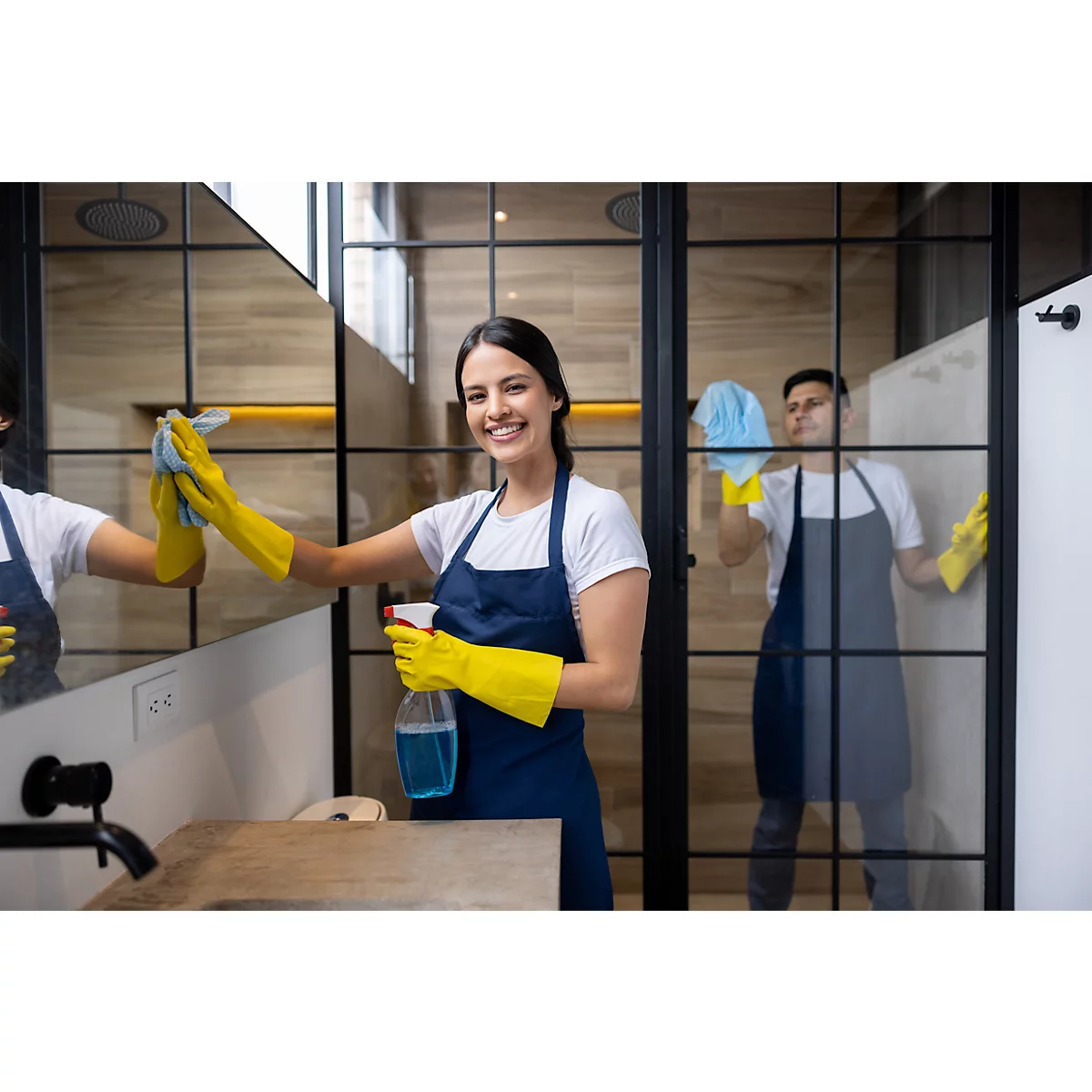Une femme nettoie un miroir dans une salle de bain. Elle porte des gants jaunes et un tablier bleu. Une deuxième personne nettoie à l'arrière-plan.