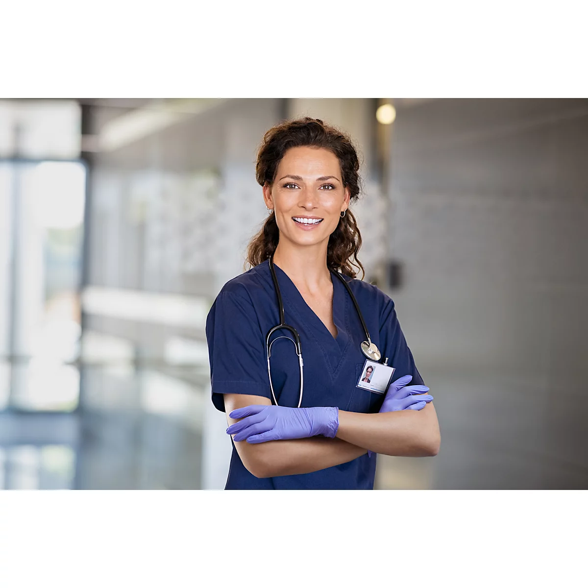 Infirmière souriante, les bras croisés dans un couloir d'hôpital. Elle porte des gants chirurgicaux et une badge nominative.