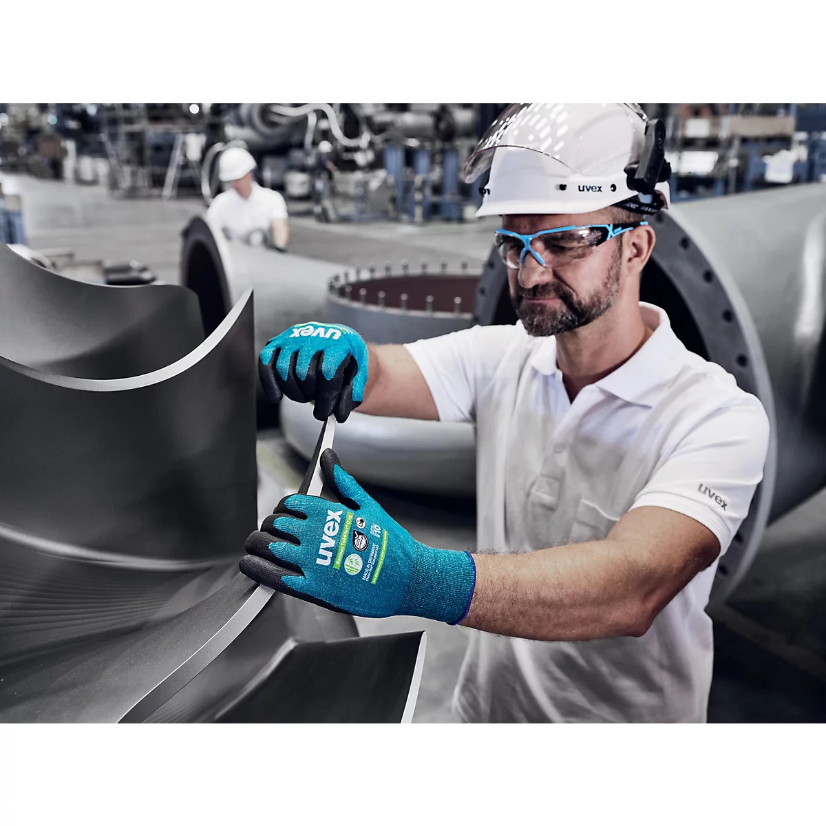 Un homme en tenue de travail examine un composant métallique. Il porte des gants bleus, des lunettes de sécurité et un casque.