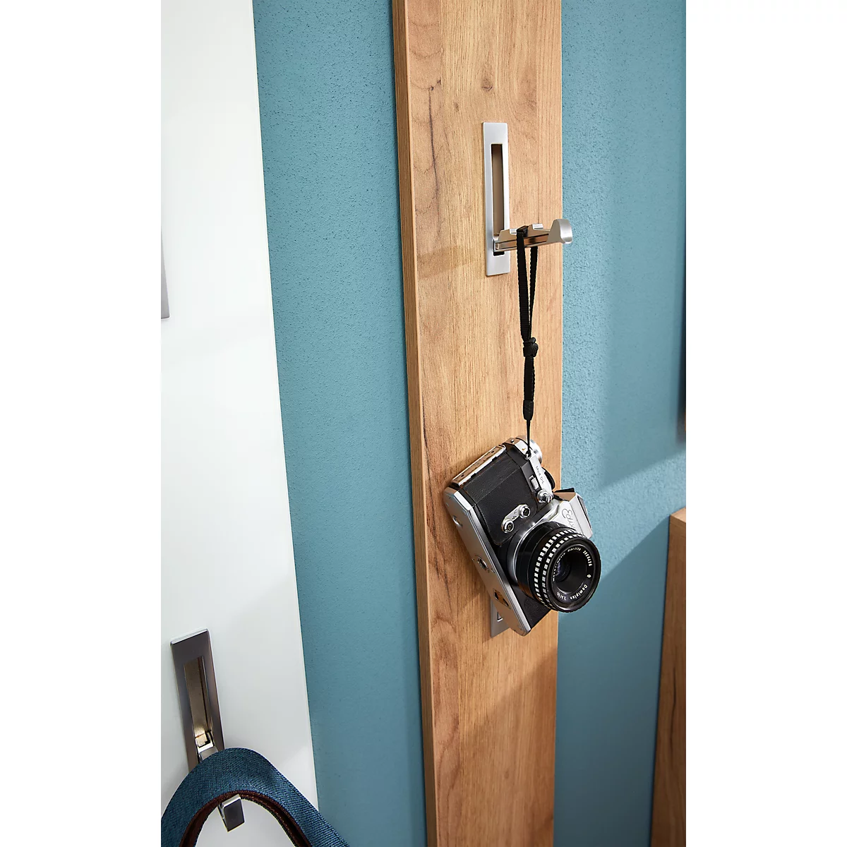 Kamera hängt an Holzlatte, die an blauer Wand befestigt ist. Haken aus Metall.