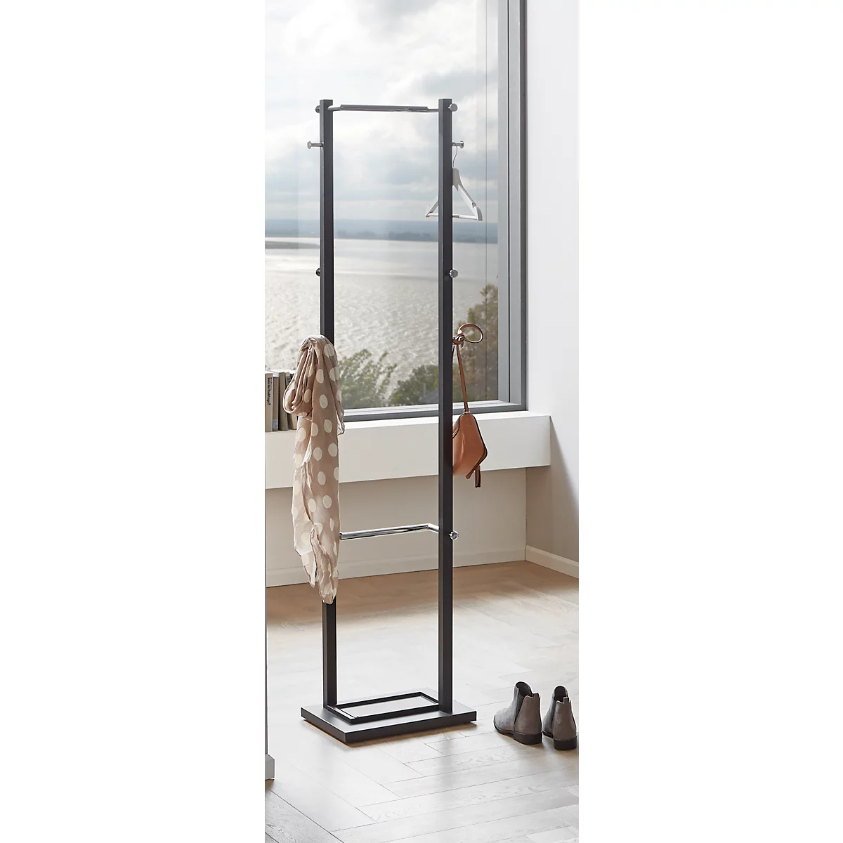 Schwarze Garderobe mit Kleiderbügeln, Schal und Tasche. Graue Schuhe stehen davor. Im Hintergrund ist eine Fensterfront mit Blick auf einen See.