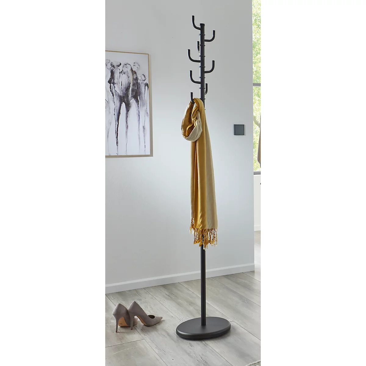 Schwarze Garderobe mit gelbem Schal, vor weisser Wand mit Kunstwerk. Schuhe liegen am Boden.