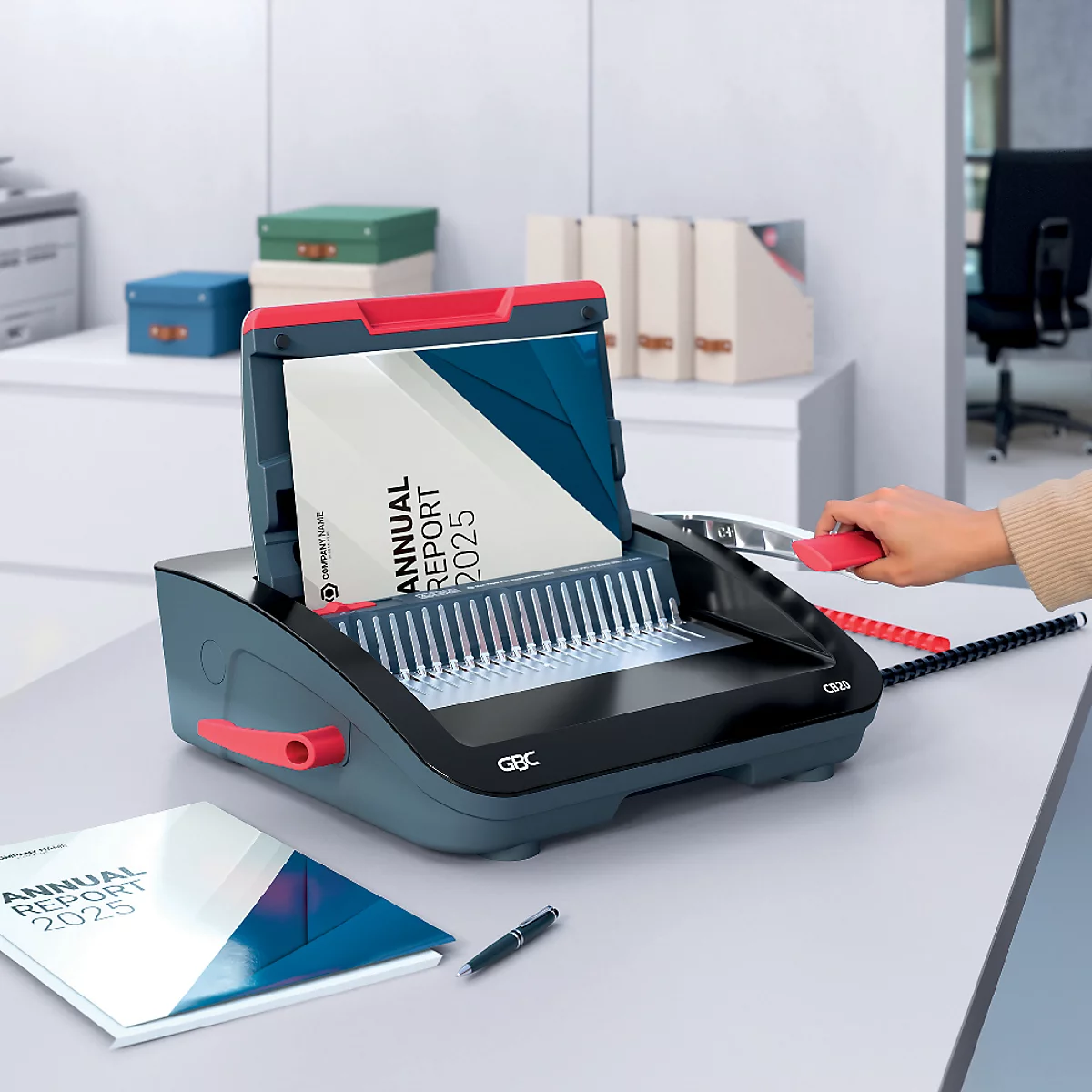 Une relieuse grise avec des accents rouges relie un rapport annuel. A côté se trouvent un rapport relié et un stylo.