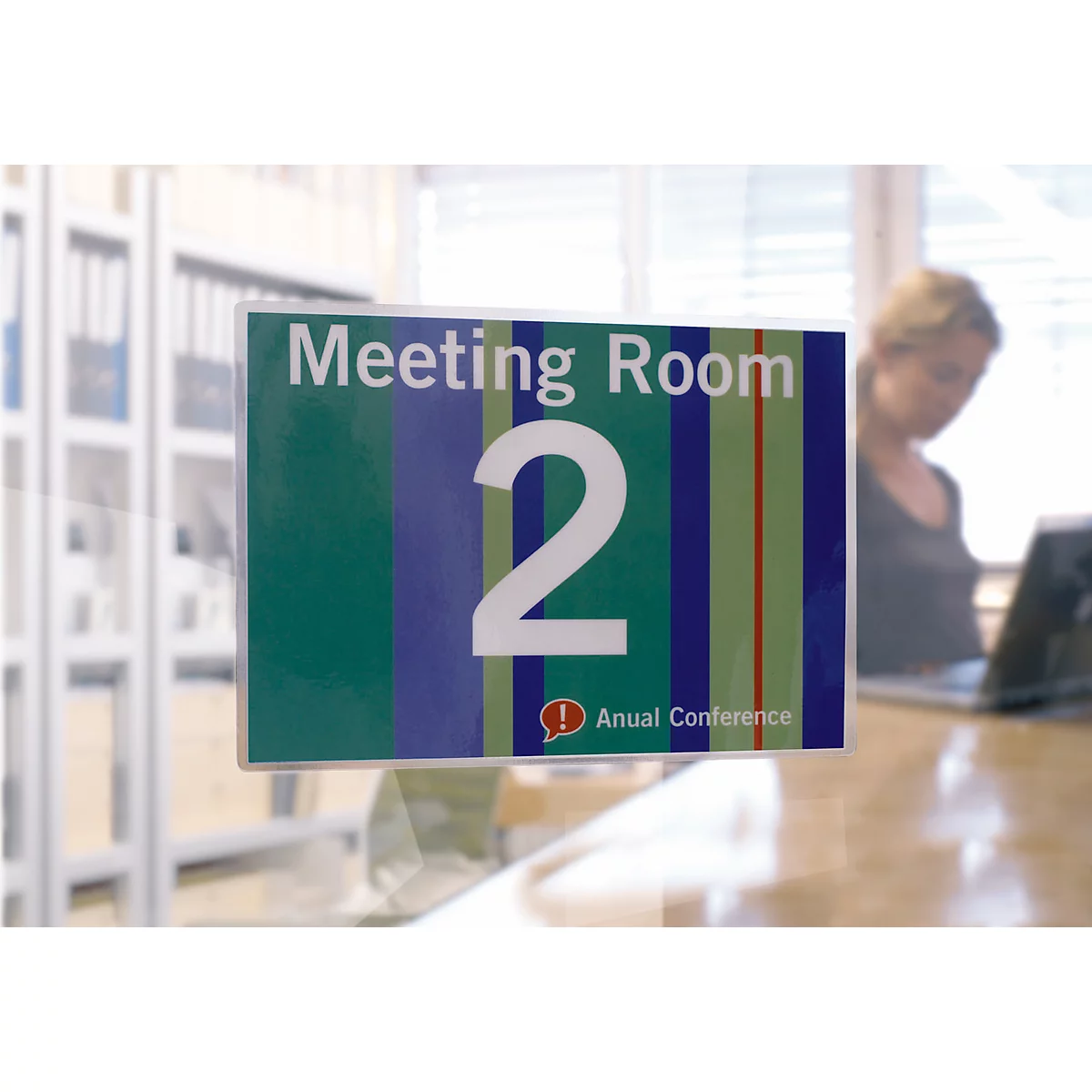 Ein Schild mit der Aufschrift "Meeting Room 2". Im Hintergrund eine Frau an einem Schreibtisch.