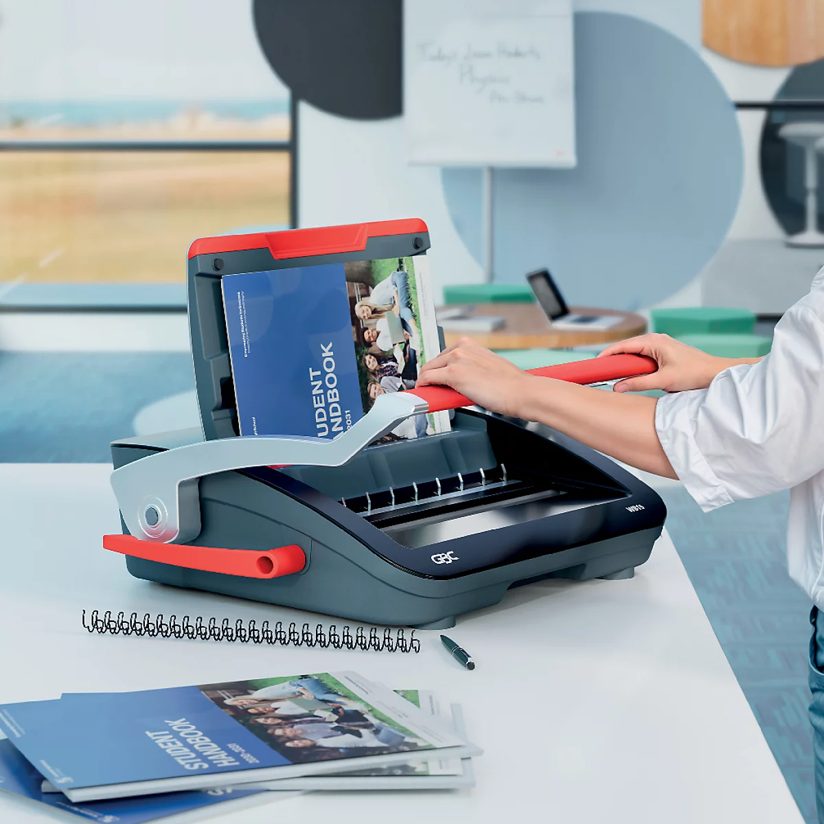 Een persoon die een boek bindt met een rode bindmachine. Er liggen nog meer boeken en pennen op de tafel.