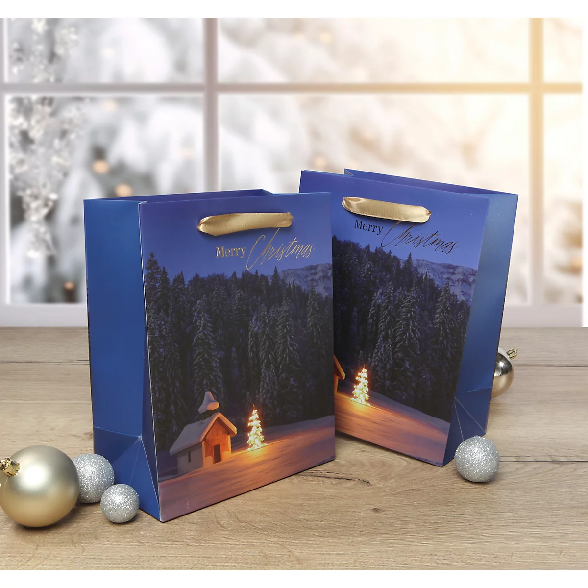 Zwei blaue Weihnachtstüten mit goldfarbenen Griffen, weihnachtlichem Motiv und der Aufschrift „Merry Christmas“ vor Winterlandschaft.