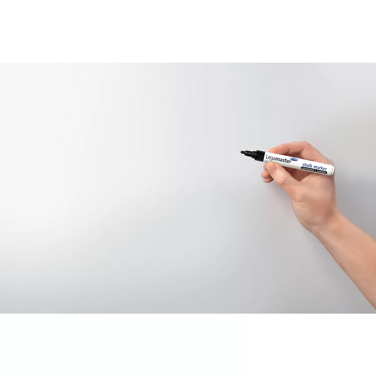 A hand holds a white marker with a black tip, ready to write on a white surface. The pen reads "Legamaster."