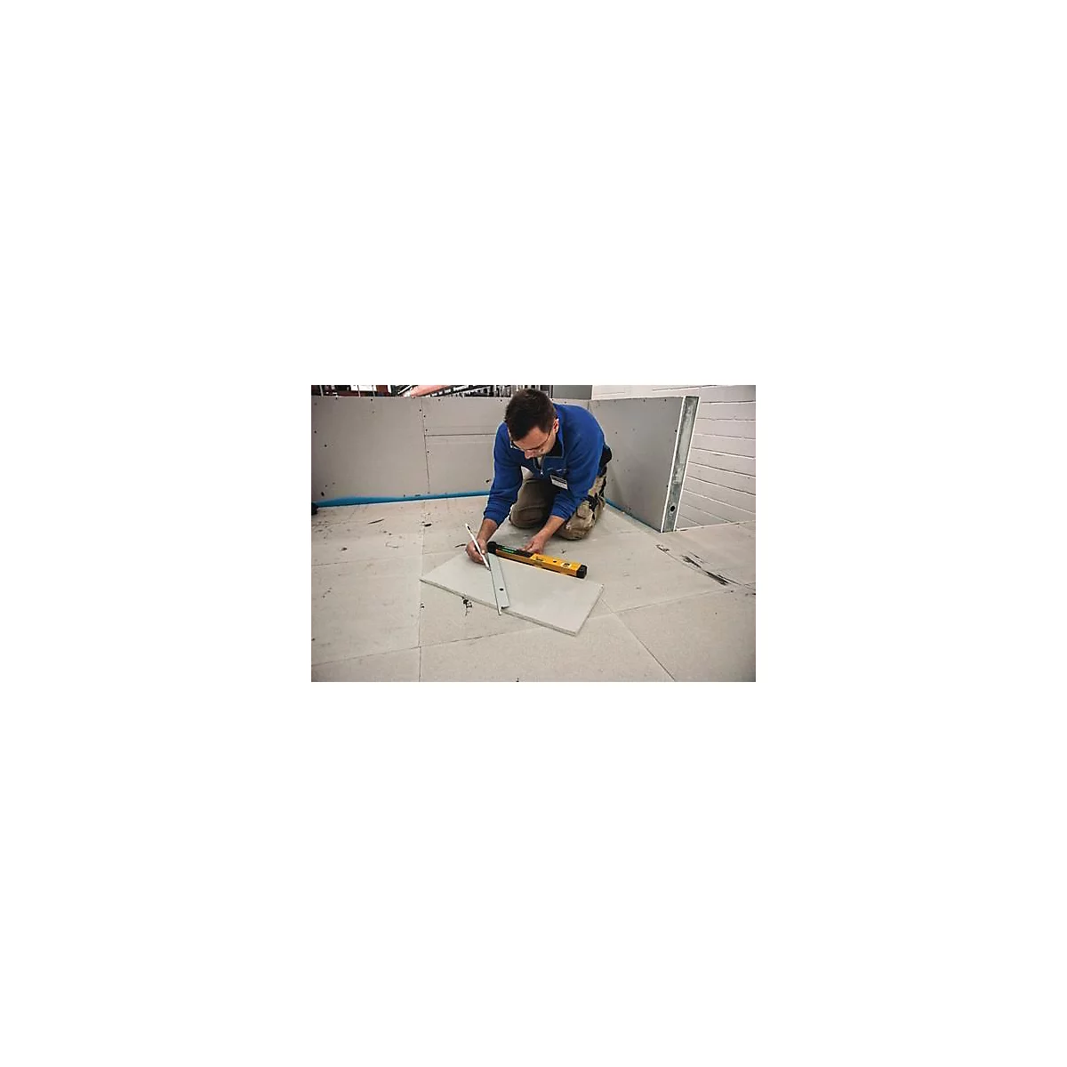 Un homme en tenue de travail bleue est agenouillé sur un sol et trace une ligne sur une dalle avec un crayon. À côté de lui se trouvent un niveau à bulle et une équerre.