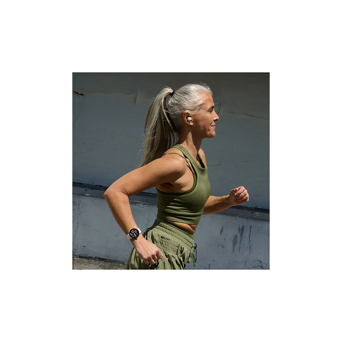 Eine Frau mit grauen Haaren joggt mit Kopfhörern und Smartwatch. Sie trägt Sportkleidung.
