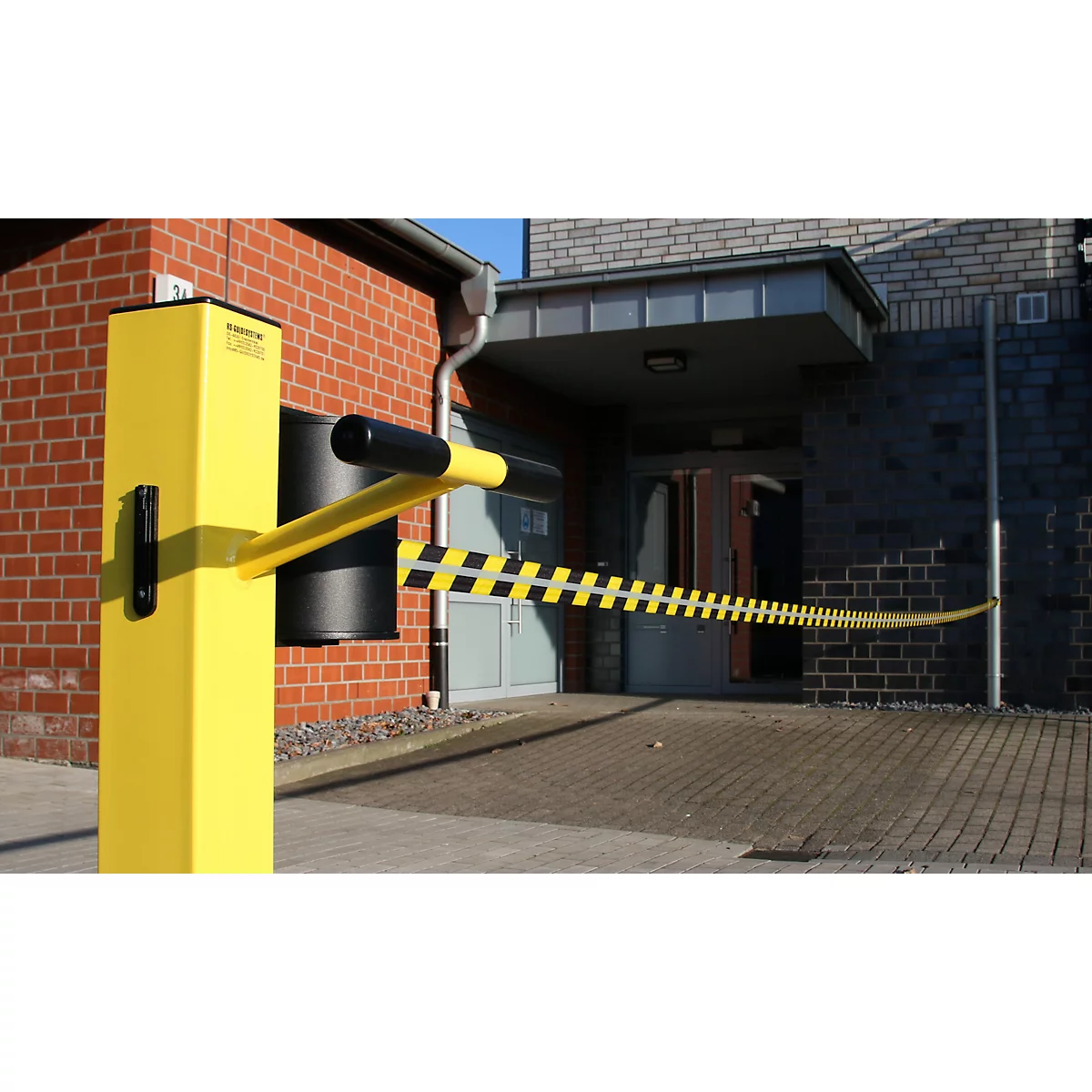 Gele slagboom met zwart-gele strepen, voor een gebouw met een rode bakstenen muur en entree.