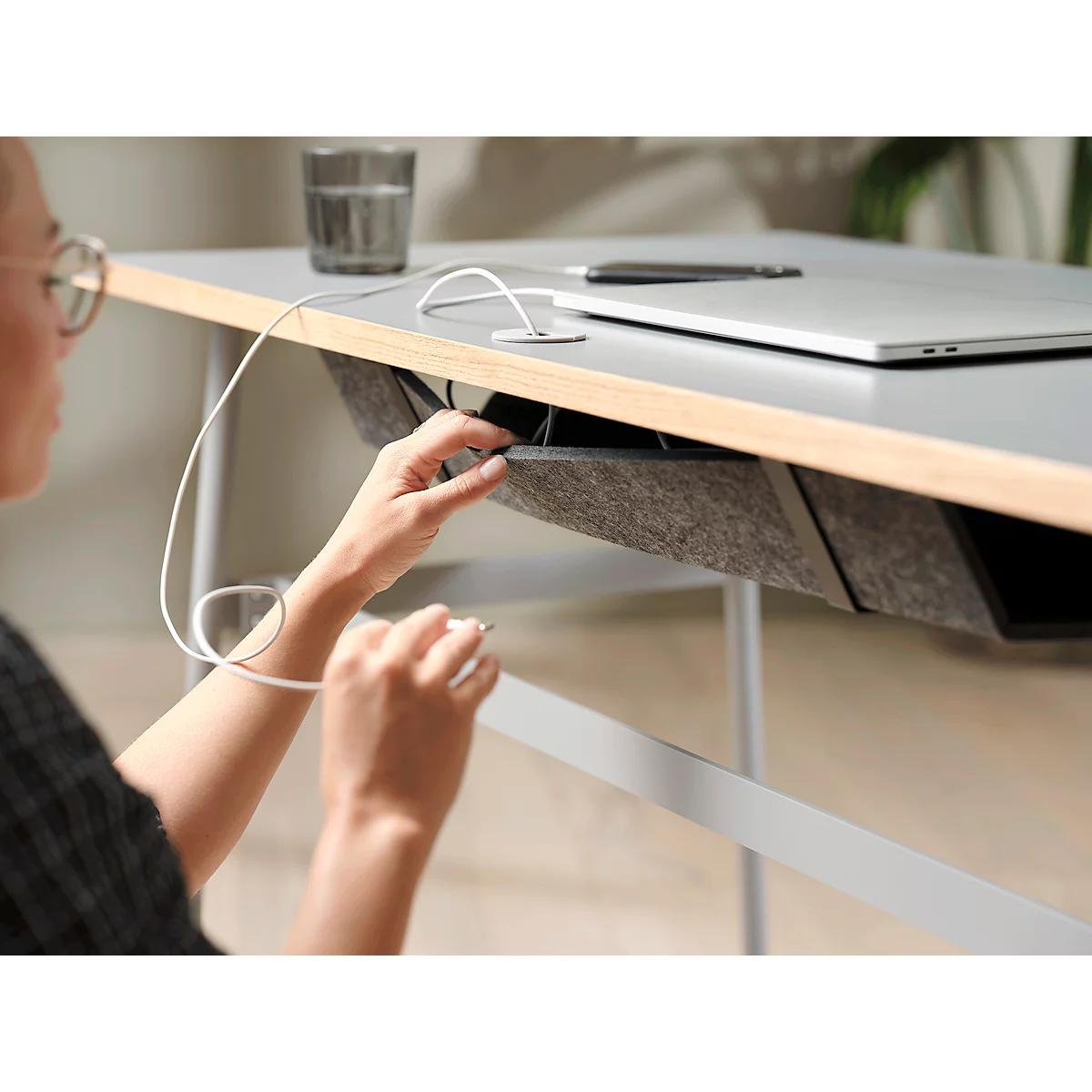 Personne avec des lunettes triant des câbles sur un bureau. Table grise avec bordure en bois, rangement en feutre, ordinateur portable.