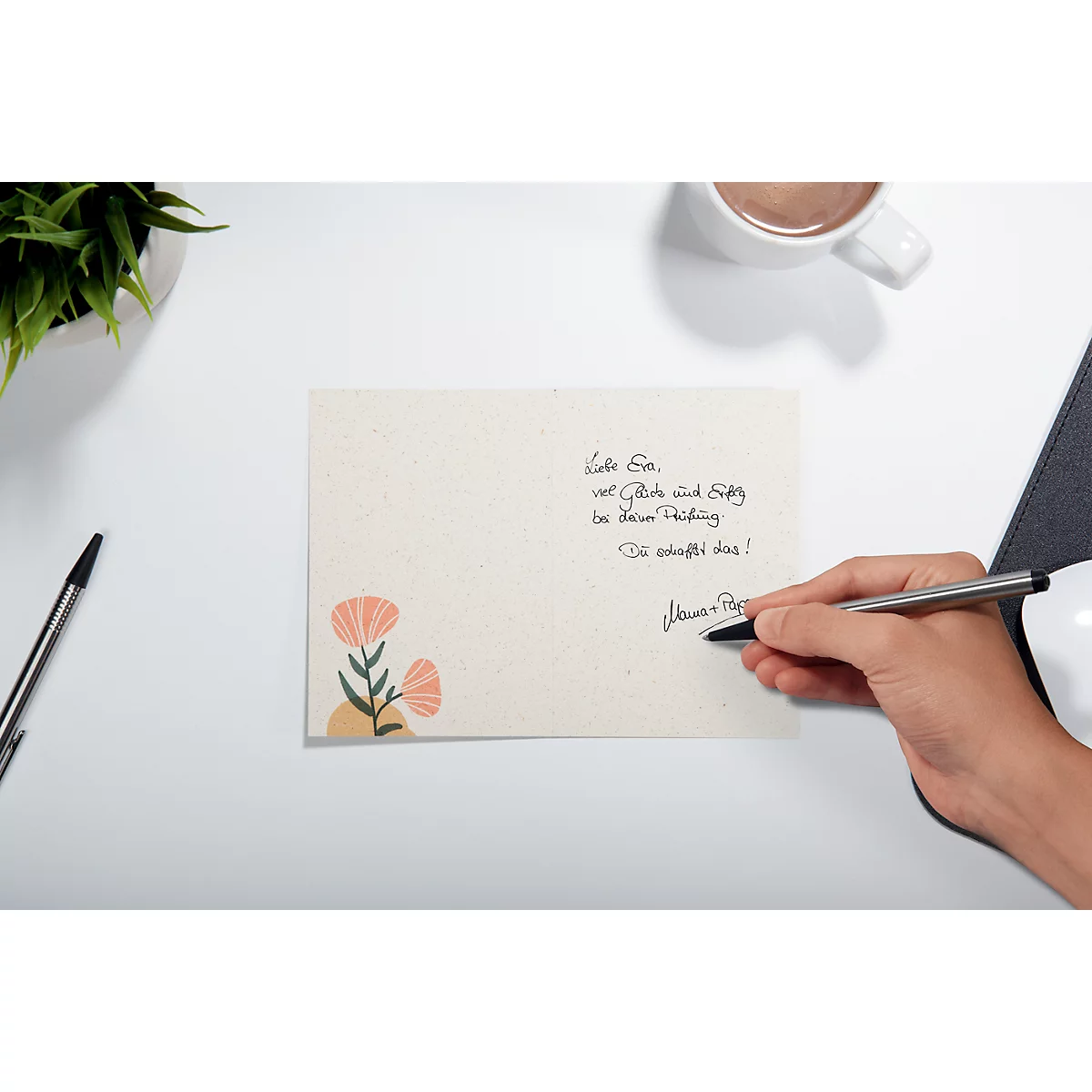 A hand writes with a pen on a card. On the card is a handwritten text, decorated with flowers.