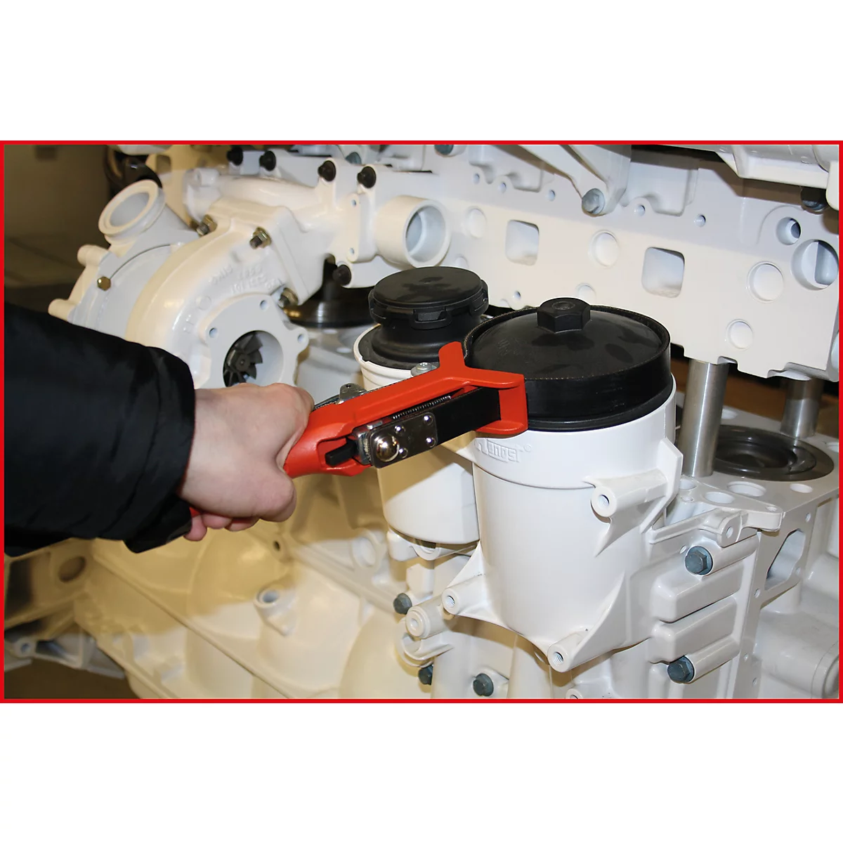 Ein Mechaniker hält einen roten Ölfilterschlüssel. Er arbeitet an einem weißen Motor. Rechts befindet sich ein schwarzer Ölfilter.