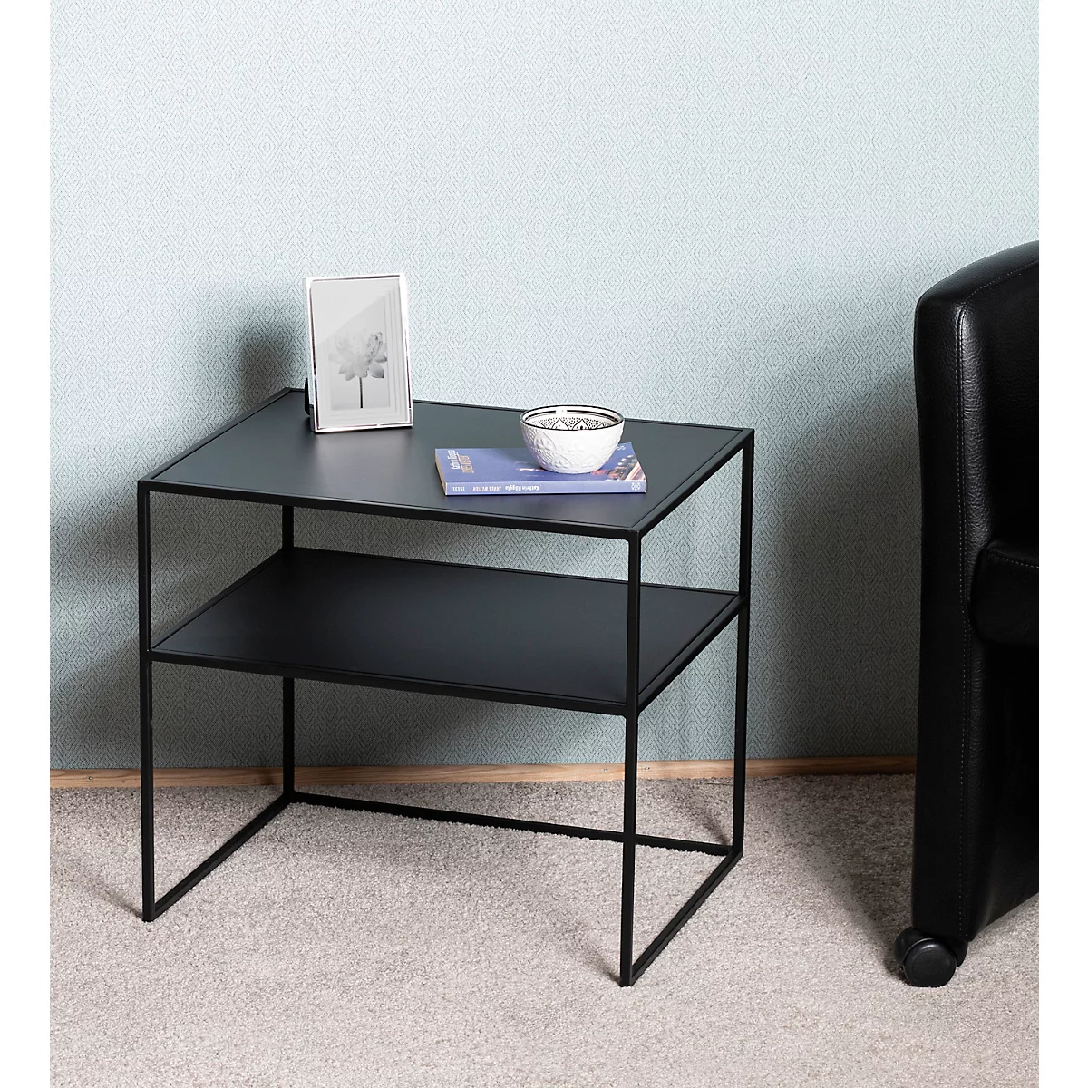 Zwarte bijzettafel met twee planken. Bovenop: foto, kom, boek. Staat voor blauwe muur, naast zwarte fauteuil.
