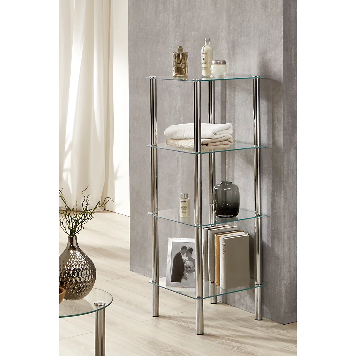 Een drielaags glazen plank met een verchroomd metalen frame. Het staat voor een grijze muur en is gevuld met handdoeken, toiletartikelen en decoratie.