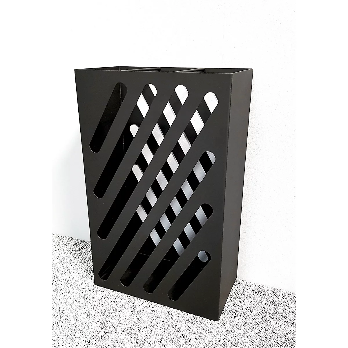 Schwarzer Metall-Mülleimer mit schrägen Schlitzen. Der Hintergrund ist weiß, der Boden grau.