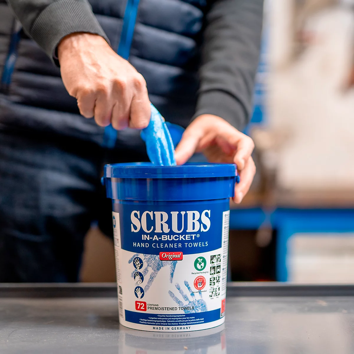 A person is taking a blue cleaning towel out of a blue bucket. The bucket says: SCRUBS IN-A-BUCKET HAND CLEANER TOWELS.