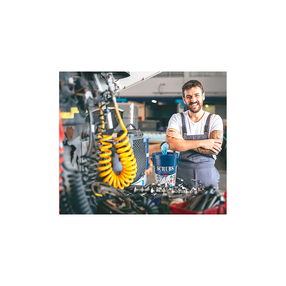 A mechanic stands in a workshop in front of a bucket of cleaning wipes, arms crossed. He smiles at the camera.