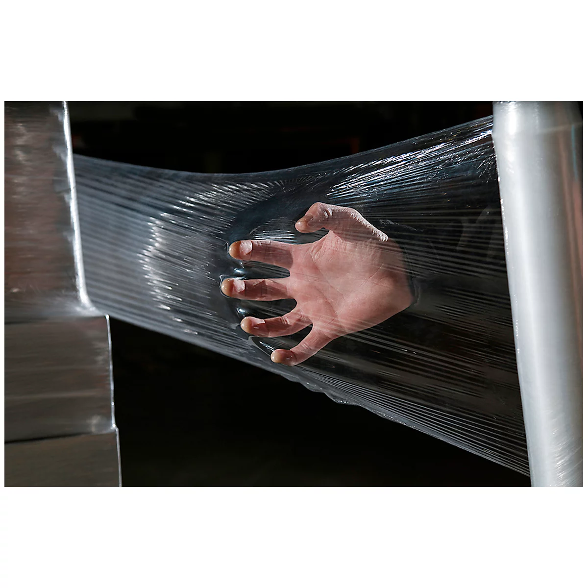 A hand presses against transparent stretch film, with a stack of pallets beneath. The palm and fingers are clearly visible, enveloped by the film.