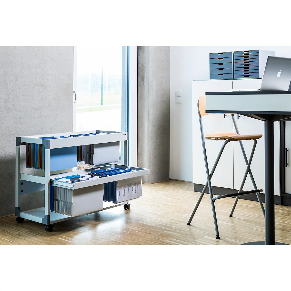 White filing cabinet on wheels, filled with folders and documents. In the background, a table with a chair and laptop.