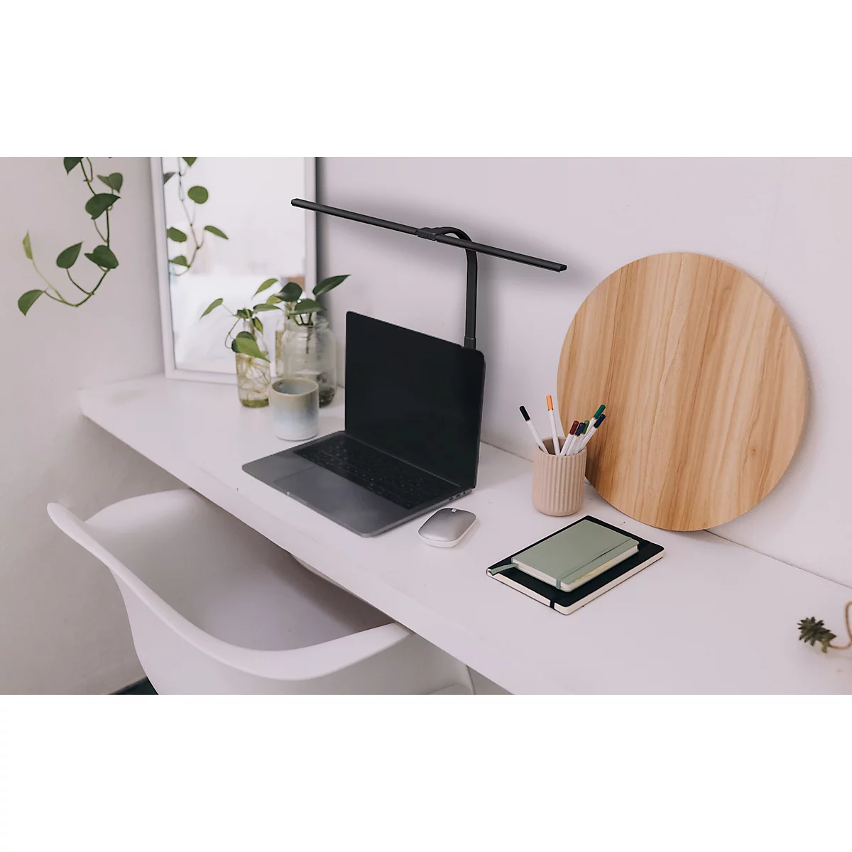 Schreibtisch mit Laptop, Lampe, Holzbrett und Stiften. Im Hintergrund ein Spiegel und Topfpflanzen.