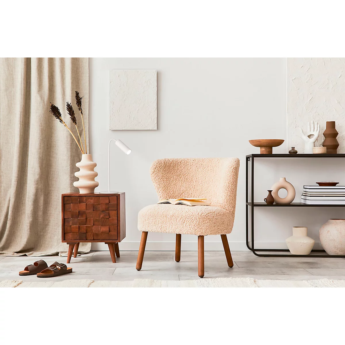 Gezellige woonkamer met crèmekleurige fauteuil, nachtkastje met vaas en boek. Een plank met decoratieve objecten, helder licht.