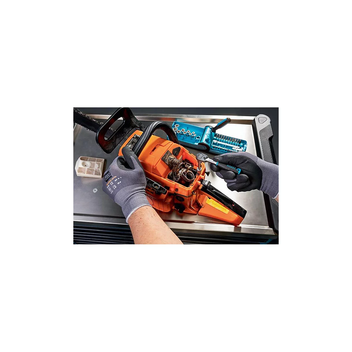 Een persoon repareert een oranje kettingzaag. Ze dragen handschoenen en houden gereedschap vast. Er liggen gereedschappen op de plank.