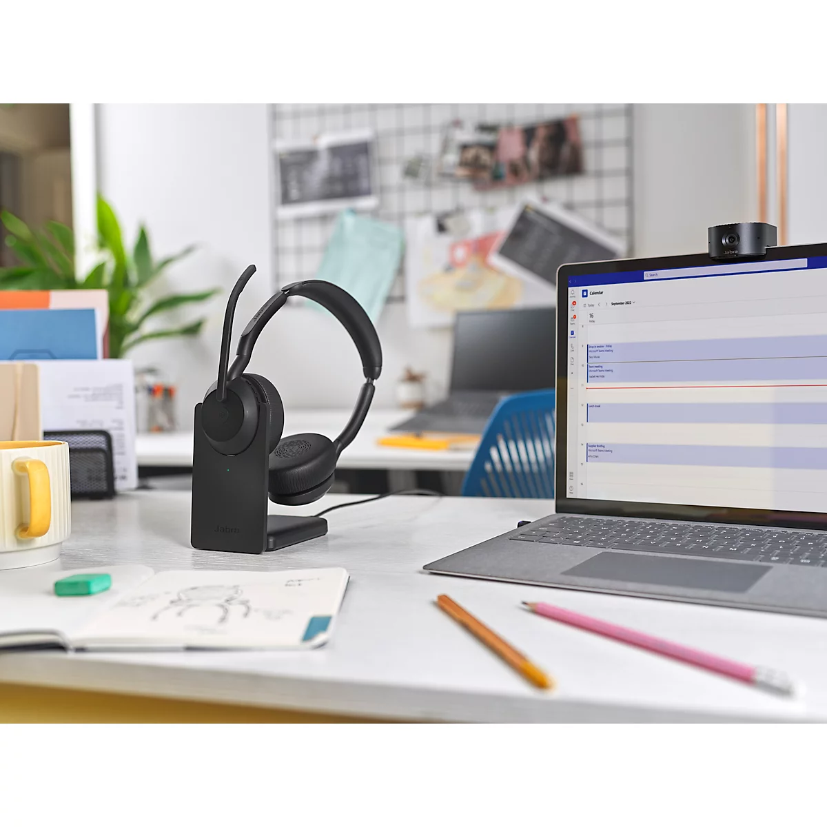 Black headset on a charging station, a laptop and other office accessories on a desk.