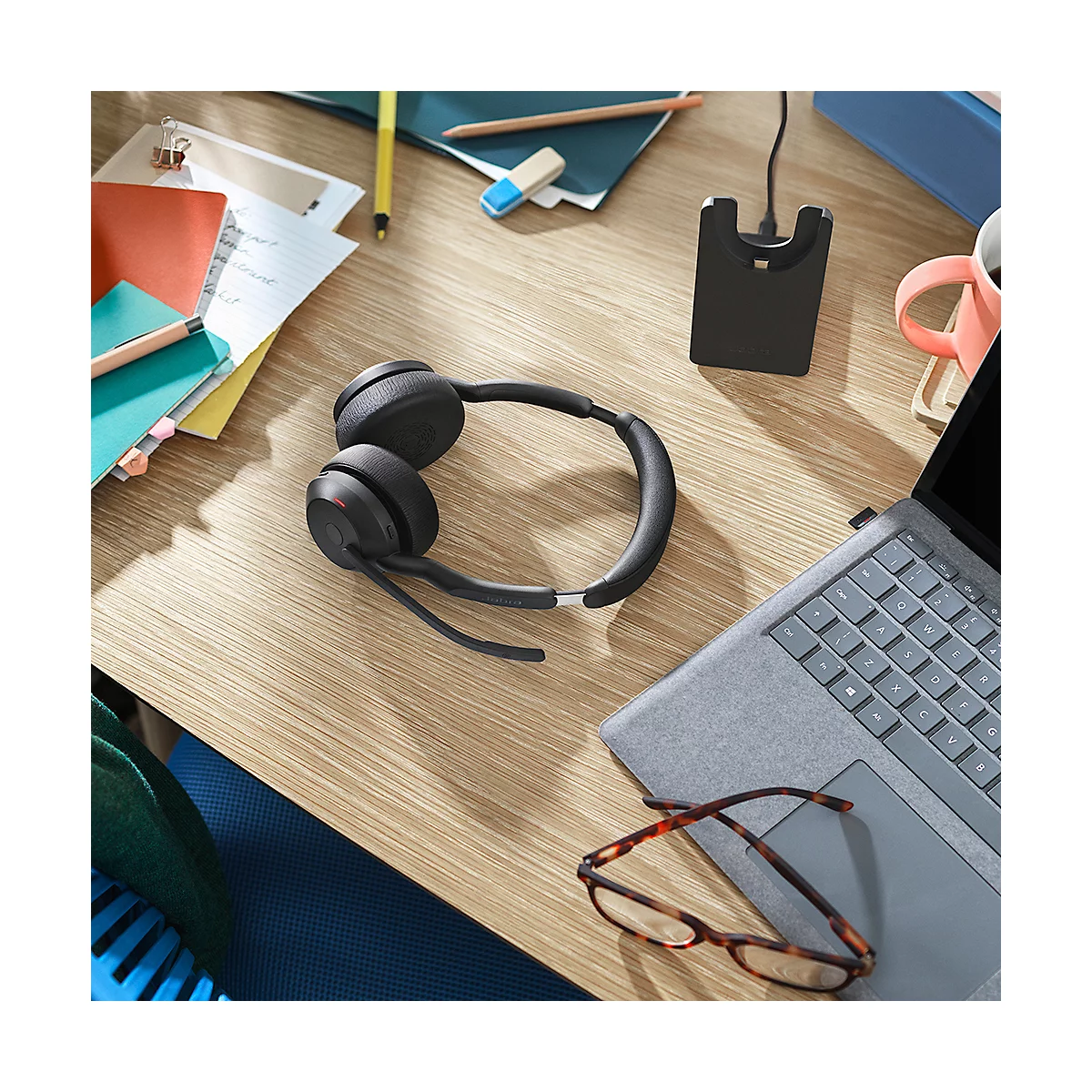 Black headset on a wooden desk, next to a laptop and glasses. In the background notes and a charging station.