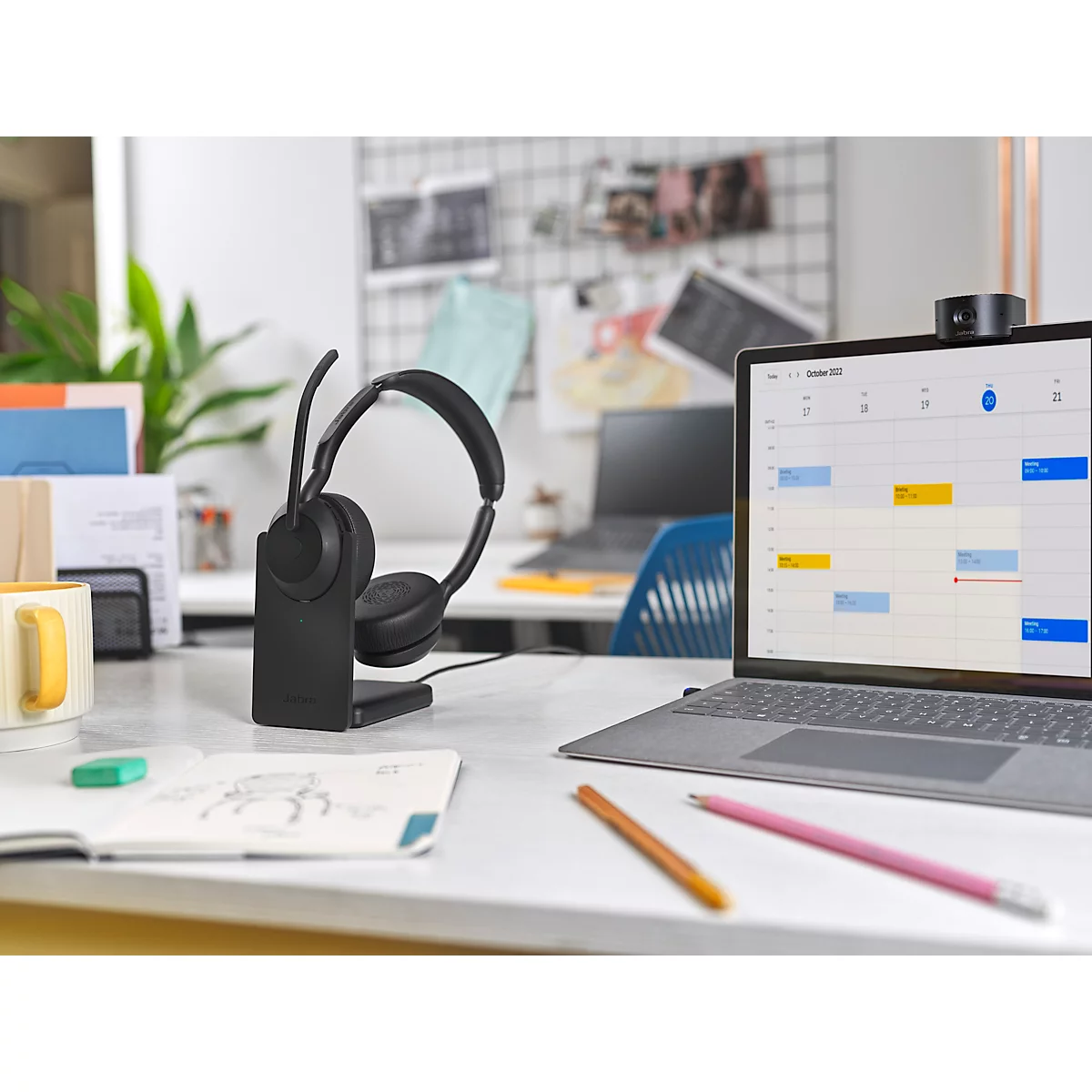 Black headset on a charging stand in front of a laptop showing a calendar. Pens and notes are on the table.