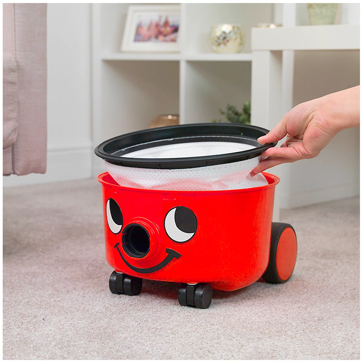 Red Henry vacuum cleaner with filter. A hand is inserting the filter. On grey carpet, background shelf.