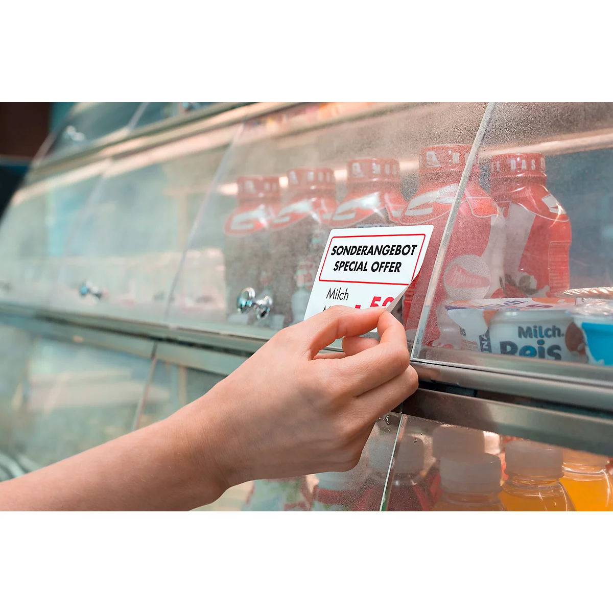 Eine Hand greift nach einem Sonderangebotsschild vor Getränken in einem Kühlschrank. Auf dem Schild steht 'Sonderangebot/Special Offer'.