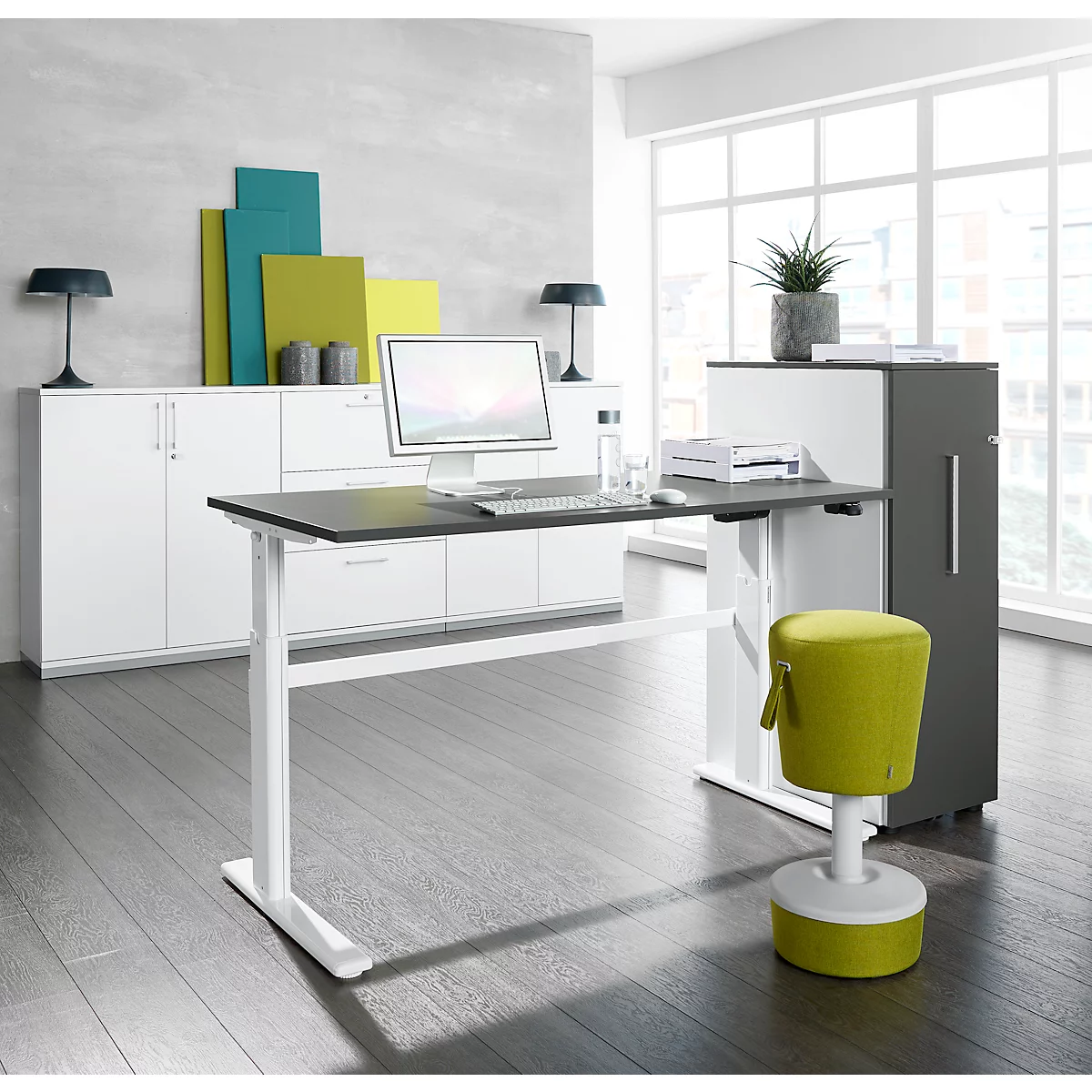 Desk with black top, white frame and green stool in front of a window front. Cabinets in the background.