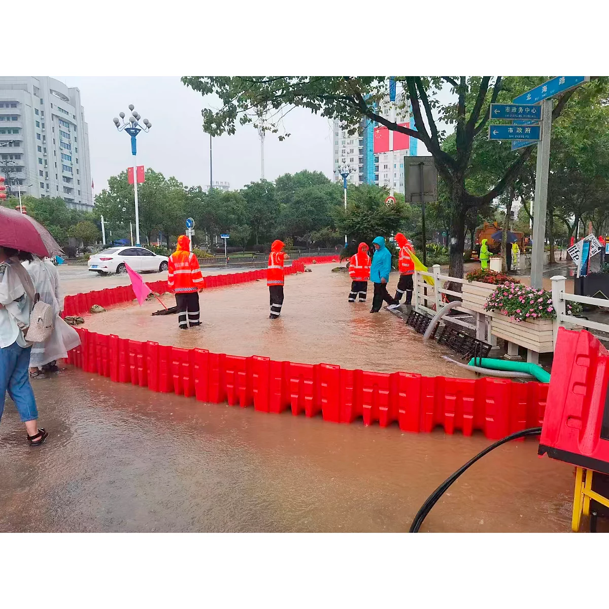 Menschen stehen in überflutetem Bereich. Rote Barrieren schützen vor Wasser. Helfer in orangefarbener Kleidung.