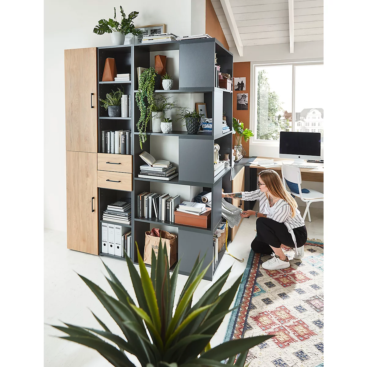 A woman in a modern home office looks at a newspaper next to a large bookcase filled with books and plants.